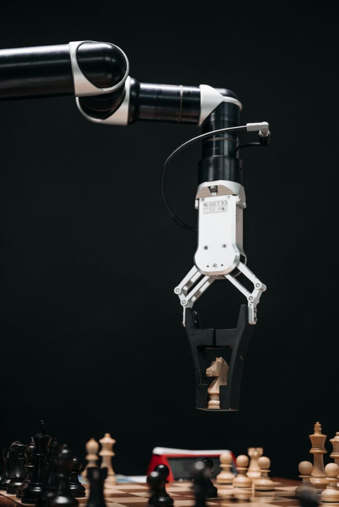 Home A robotic arm with a pincers holding a knight chess piece on a chessboard.